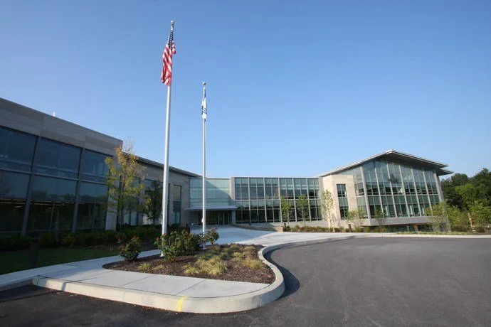 Modern office building with glass windows, two flagpoles with American flags, a curved driveway, and landscaped surroundings.
