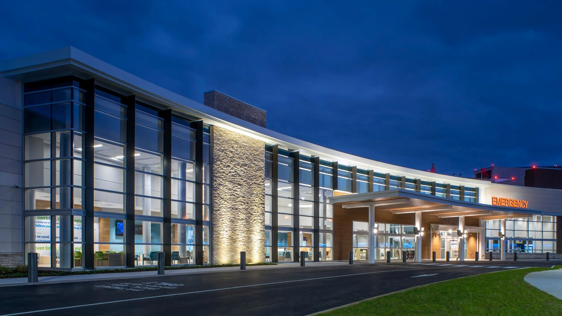 Modern hospital emergency entrance with glass facade, illuminated signs, and parking area at night.