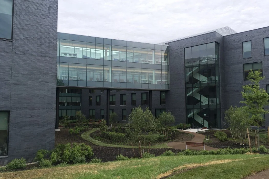 Modern multi-story building with dark gray brick exterior, large glass windows, and an outdoor stairway. Surrounding landscaped garden with trees, bushes, and a small walking path.