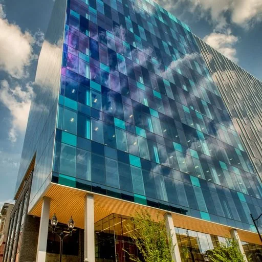 Modern glass office building reflecting the sky and clouds, with a yellow-colored lower facade and greenery at the base.