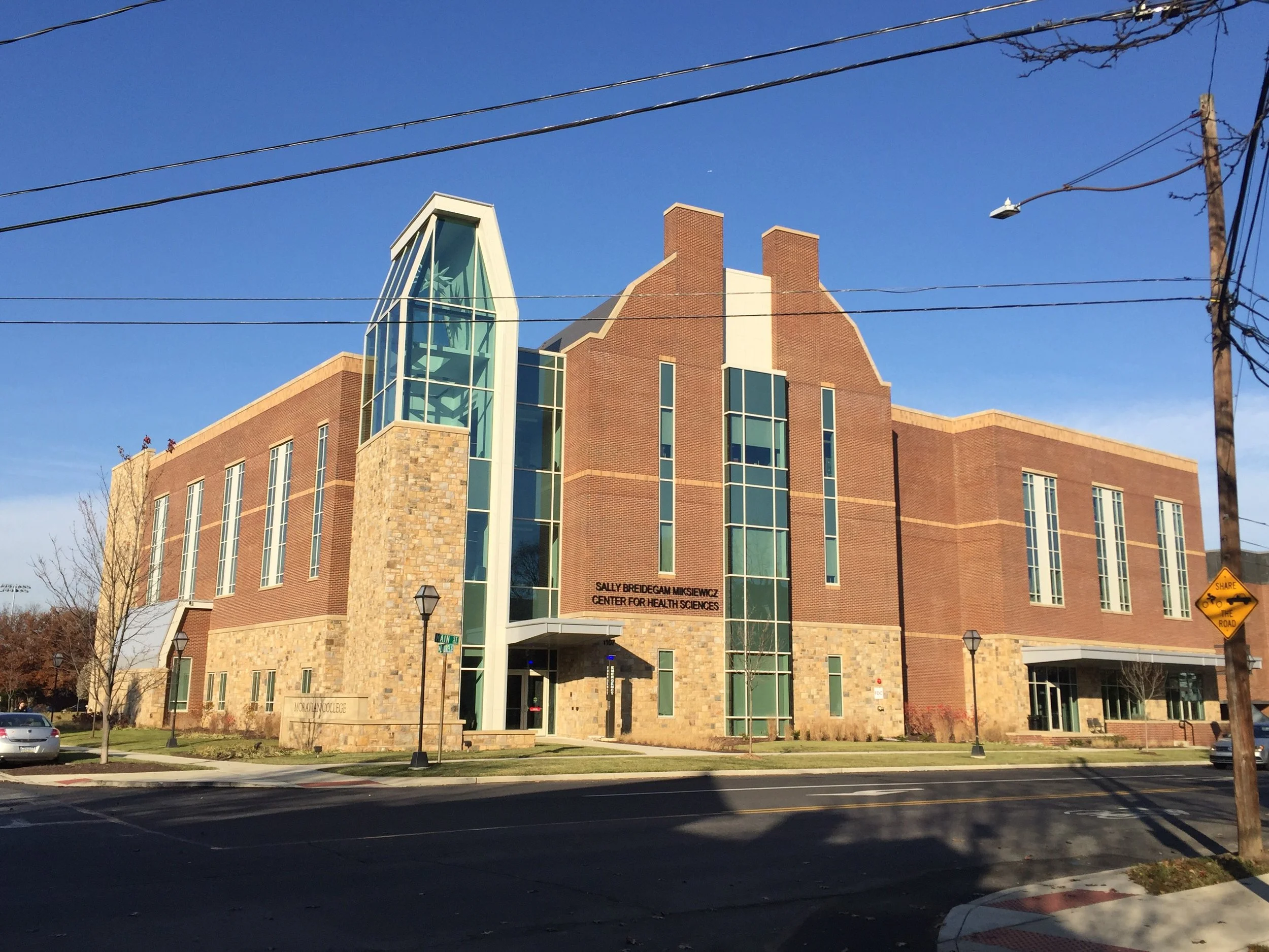 The Sally Breidegam Mikaelian Center for Health Sciences, a large brick educational building with modern glass extensions, situated on a college campus. There are a few cars parked in front and some leafless trees. A street sign indicates the intersection of Main Street and another road.