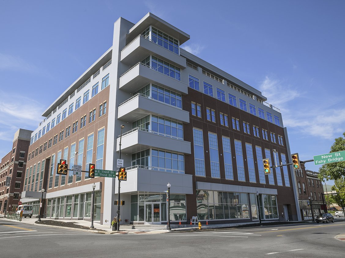 Modern multi-story building with glass windows, located at the intersection of E Third Street and New Street Fahy Bridge, with traffic lights and street signs.