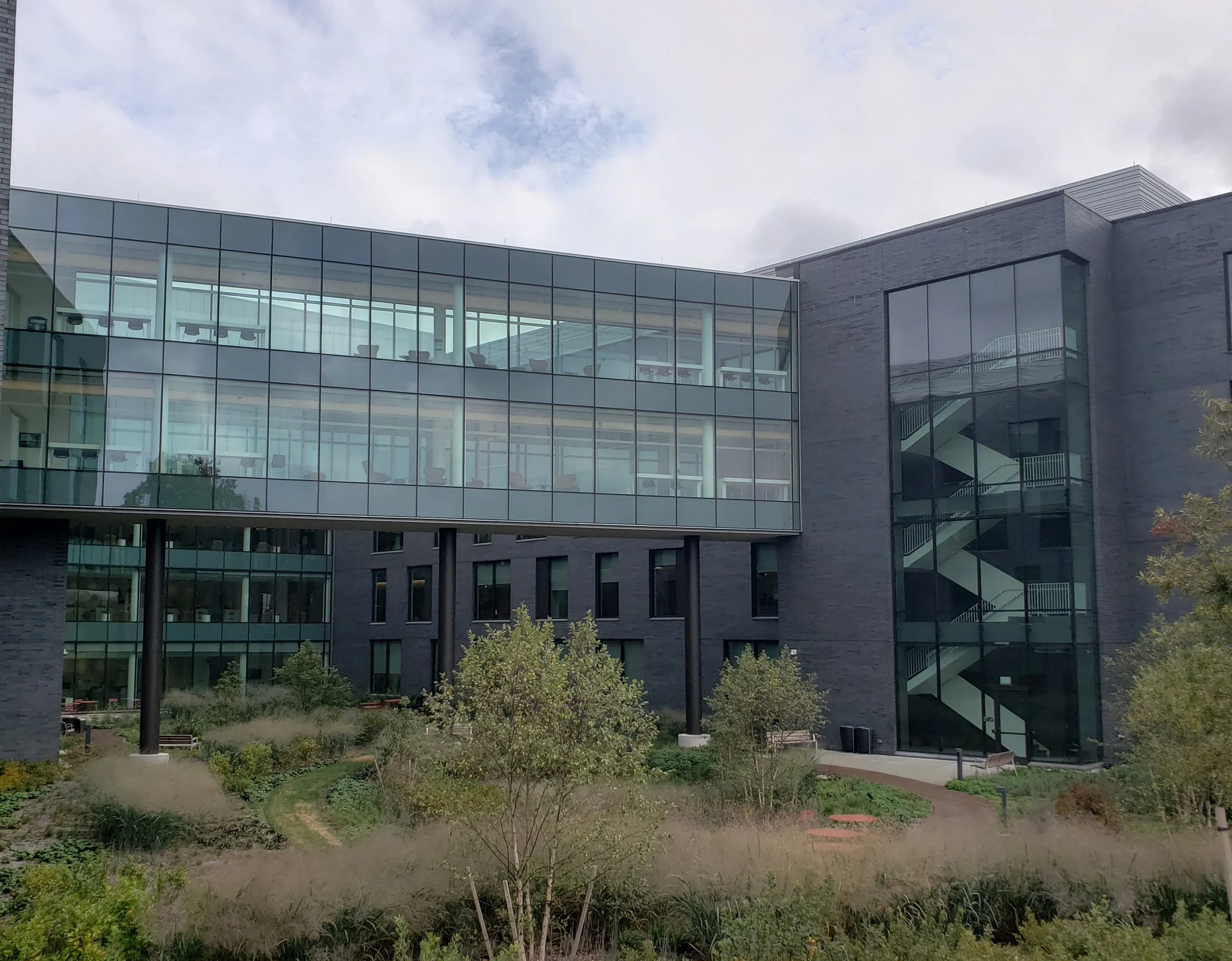 Modern building with glass windows overlooking an outdoor garden area with trees and walking paths, under a cloudy sky.