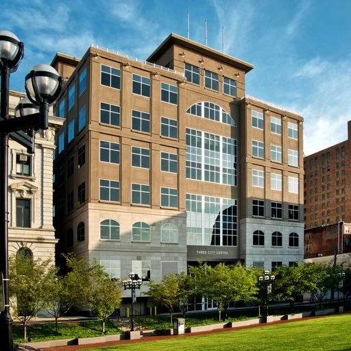 Exterior view of a modern multi-story office building with large windows, surrounded by a park with trees and lamp posts, under a blue sky.