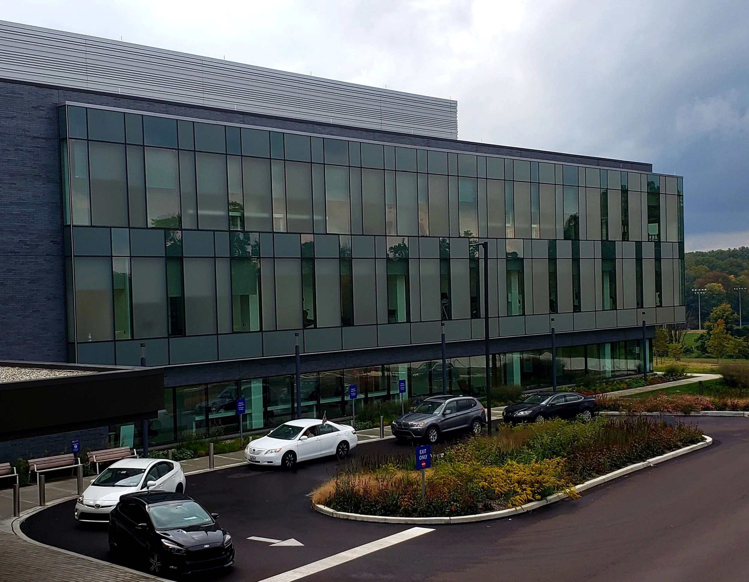 A modern building with large glass windows and horizontal metal siding on the roof. There are multiple parked cars in a parking lot with landscaped areas and trees in the background. The sky is overcast with gray clouds.