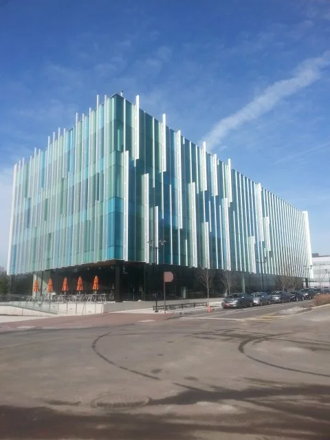 Modern multi-story building with glass and metal exterior, parked cars along the street, and a blue sky with some clouds.