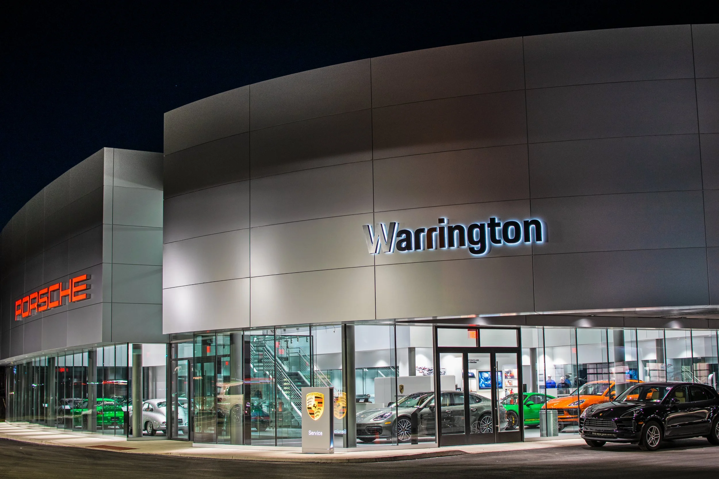 Exterior view of a luxury car dealership with Porsche and Warrington signs, displaying several sports cars inside through glass windows at night.