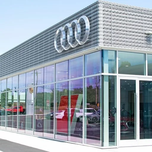 Audi car dealership with Audi logo on the building and cars displayed inside through large glass windows.