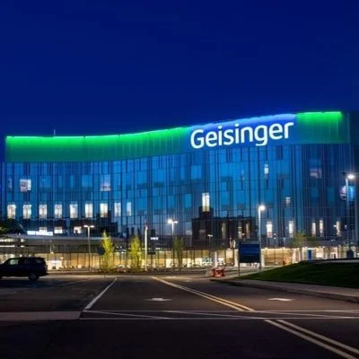 A modern building with the name 'Geisinger' lit up on the roof at night, with a parking lot and some trees in front.