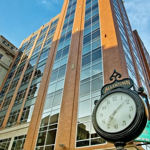 A tall brick and glass building with reflective windows and a large clock that reads approximately 7:02, labeled 'Allentown'.