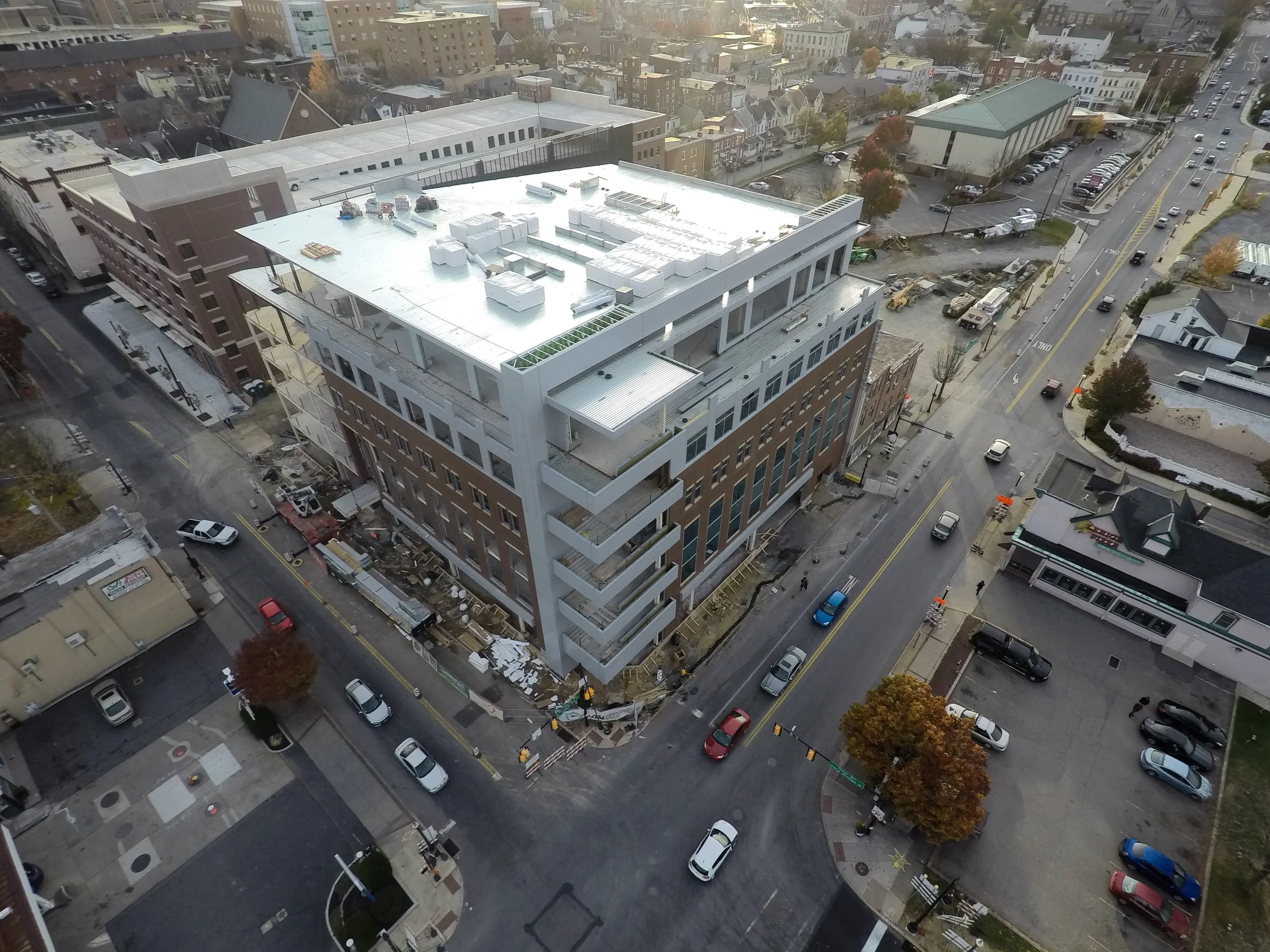 Aerial view of a city block with a large building under construction or renovation on the corner, surrounded by streets with parked cars, trees, and other smaller buildings.