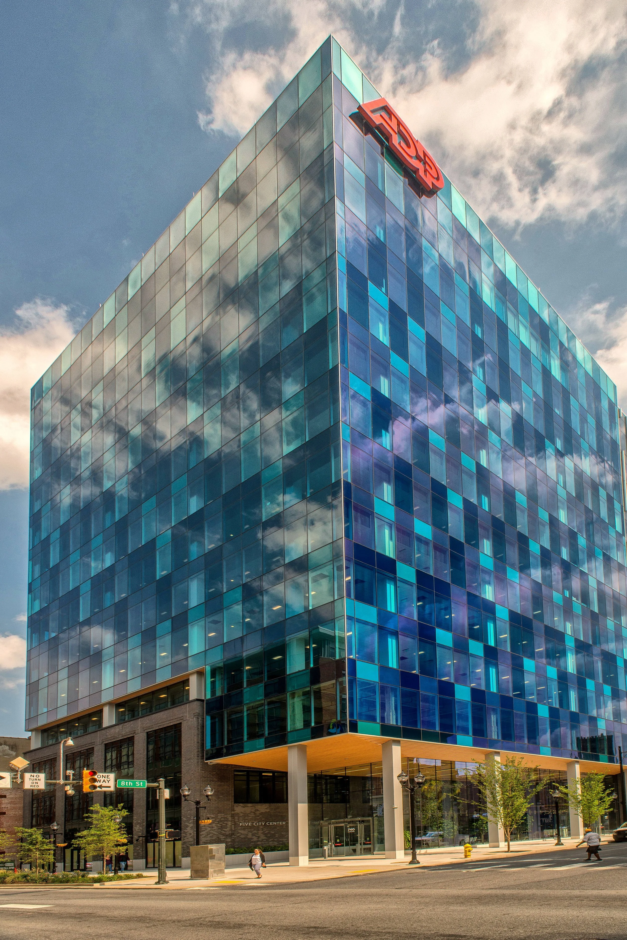 A modern glass skyscraper with blue and green tinted windows, featuring the red 'FNB' logo at the top, located at the corner of a city street with a few pedestrians, streetlights, and traffic signs.