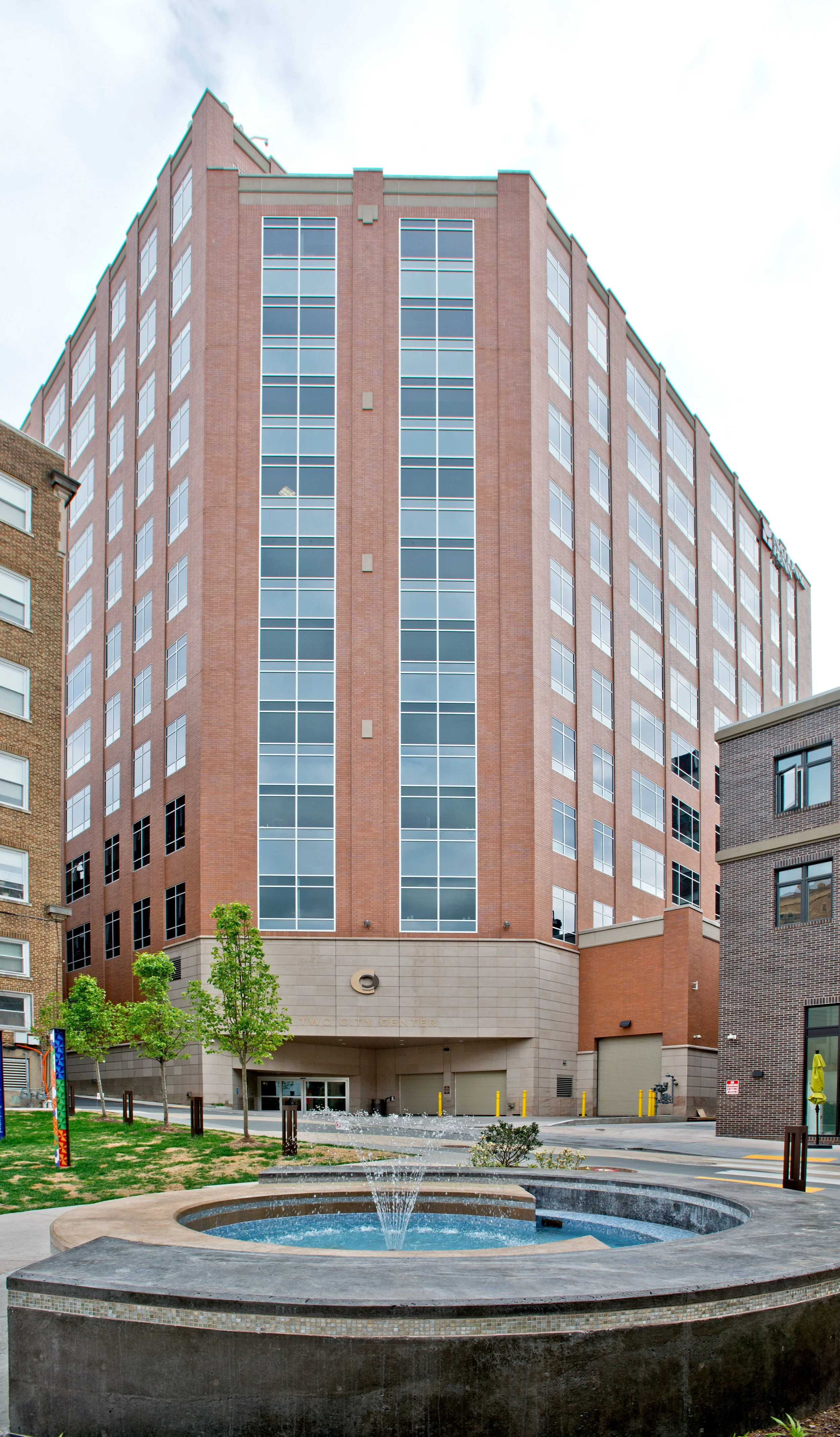 A high-rise brick building with large windows, located in an urban area with smaller buildings nearby. In the foreground, there is a small fountain surrounded by a walkway, trees, and some street furniture.
