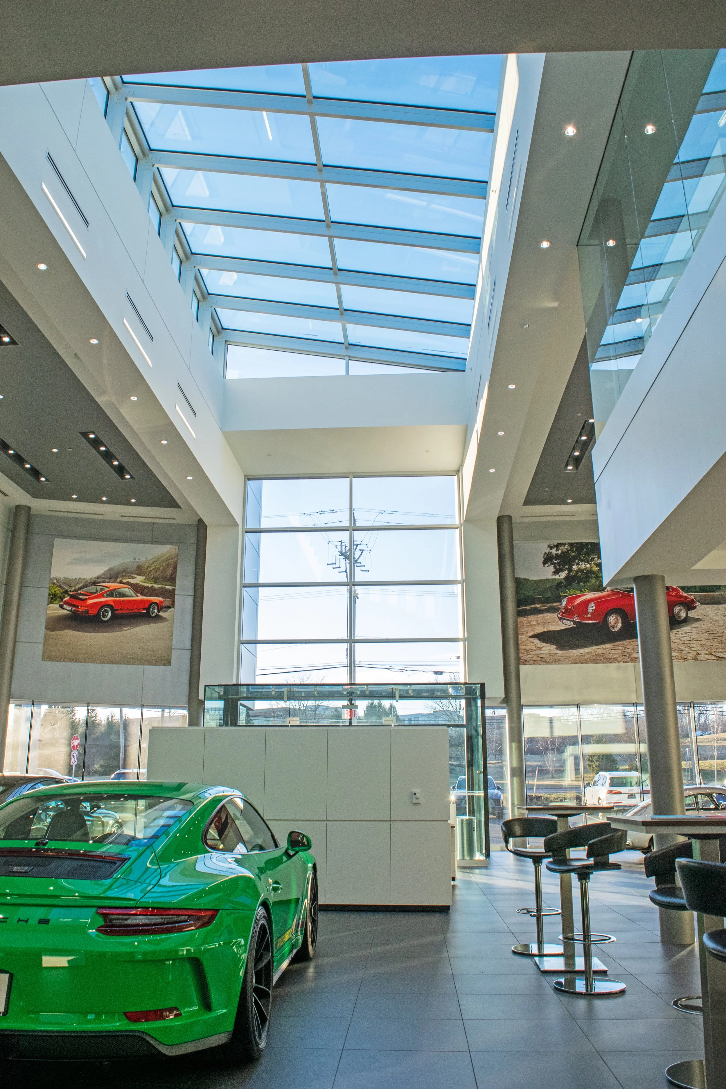 A bright, modern car dealership showroom with a green sports car in the foreground and large windows and skylights, allowing natural light to fill the space.
