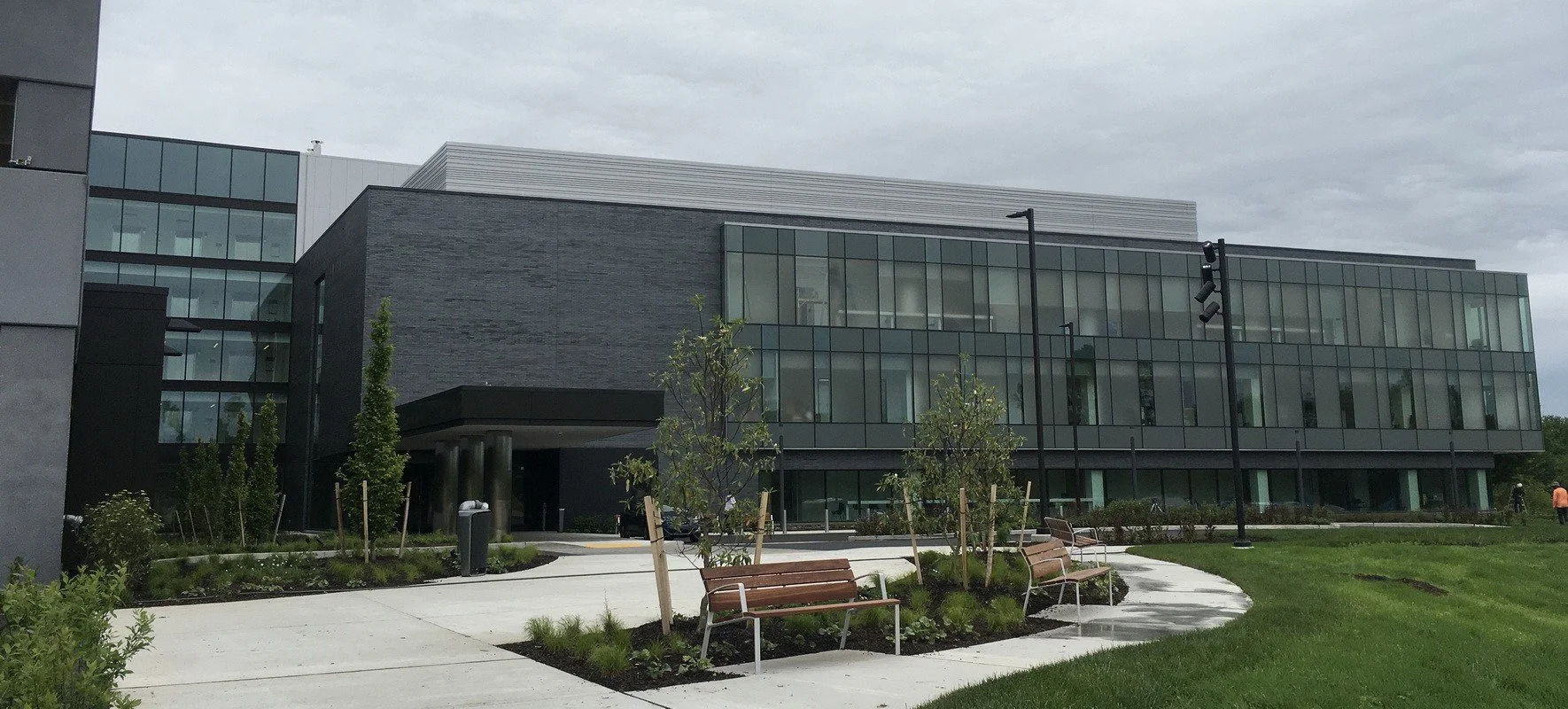 Modern multi-story building with glass windows, surrounded by small trees, benches, and an open grassy area under a cloudy sky.