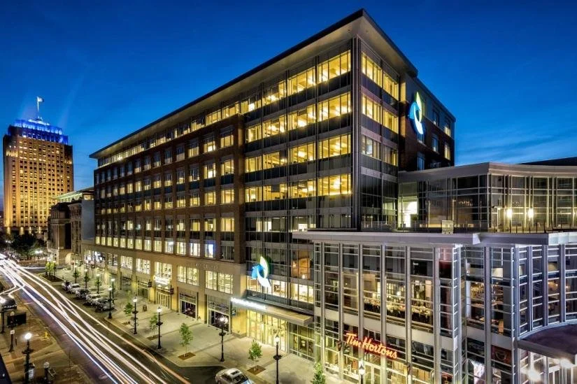 Nighttime cityscape featuring a modern glass building with yellow-lit interior, Tim Hortons storefront on the ground level, and a busy street with light trails from passing cars.