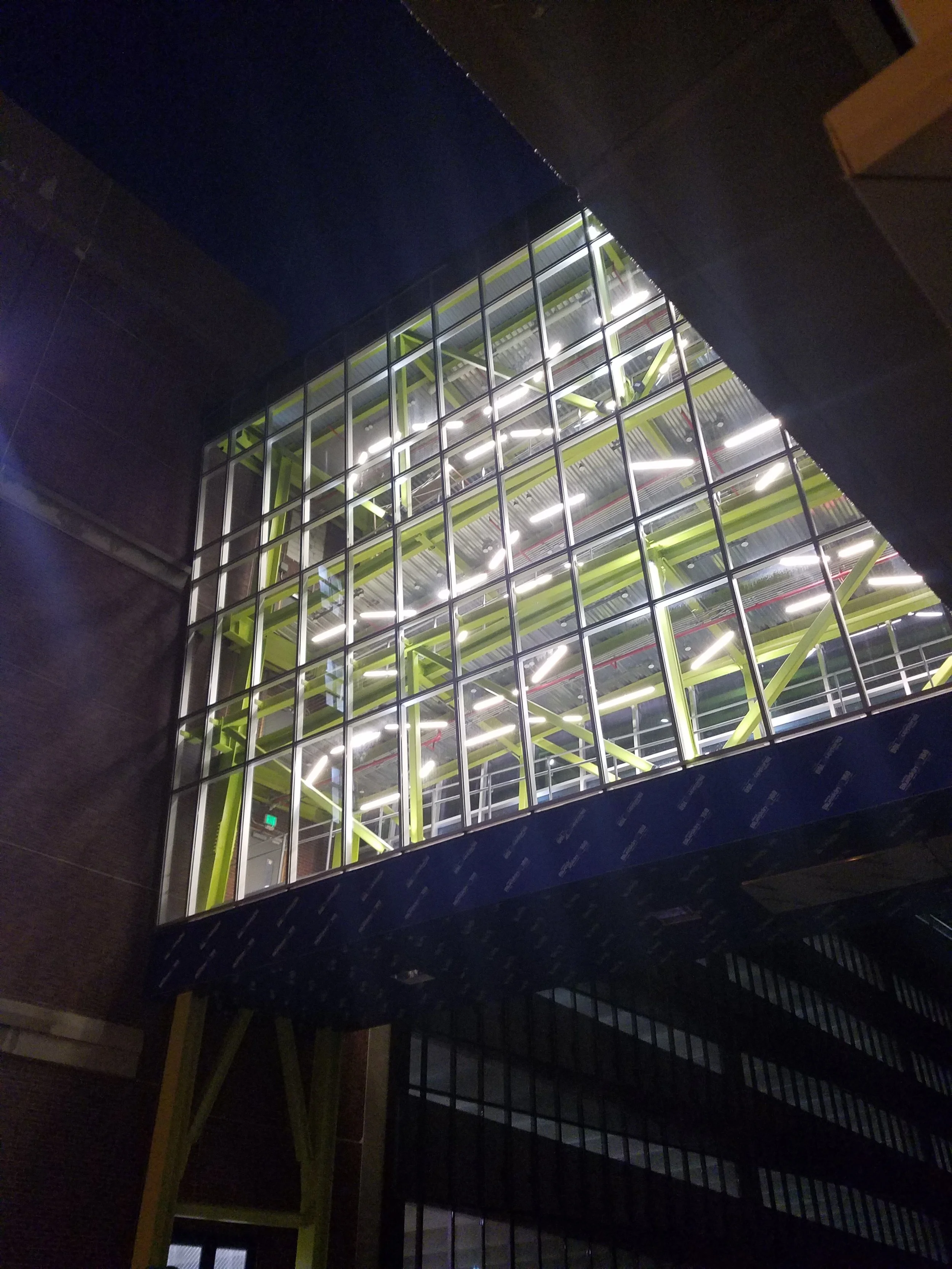 Nighttime view of a modern building with large glass windows showing an interior with green steel beams and bright lighting.