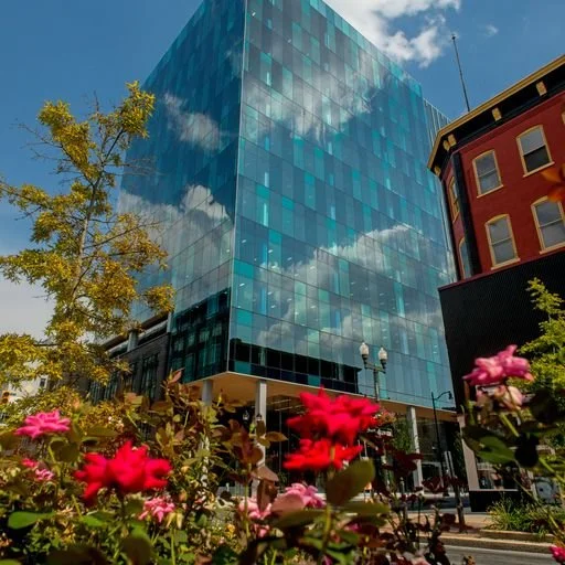 Modern glass office building reflecting a blue sky with clouds, surrounded by trees and colorful flowers in an urban setting.