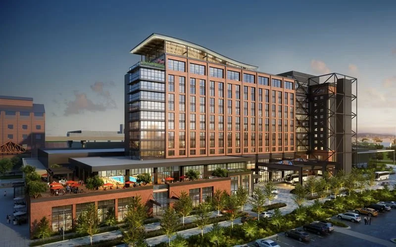 A modern multi-story hotel or office building with glass windows and a rooftop terrace, surrounded by trees and a parking lot under a cloudy sky.