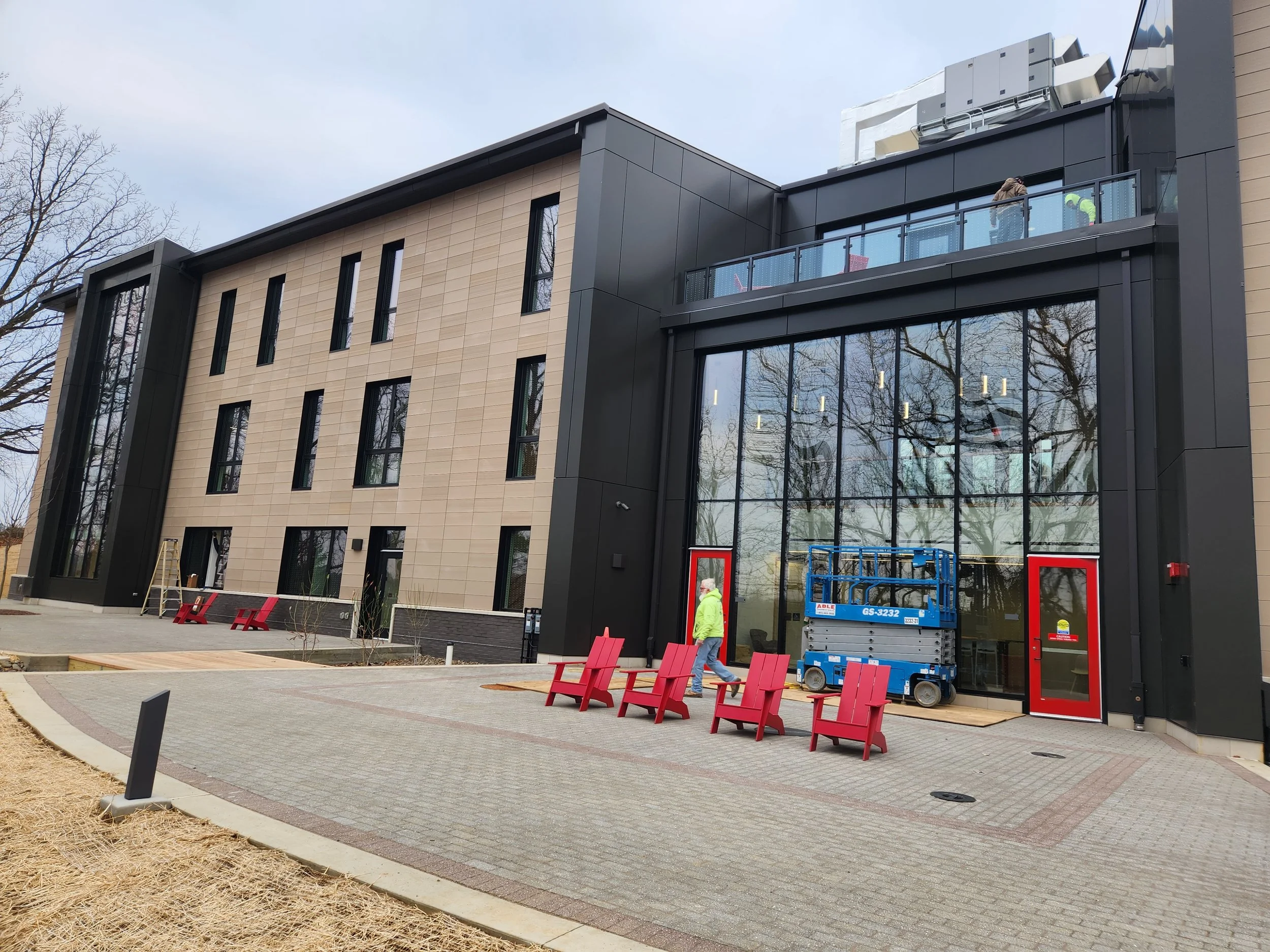 Modern building under construction with workers on a balcony, construction equipment, red benches, and a paved walkway in the foreground.