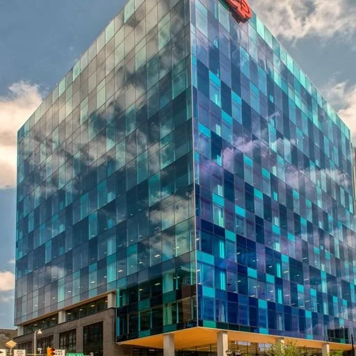 A modern glass office building with blue-tinted windows, reflecting the sky and clouds, with a red logo at the top corner.