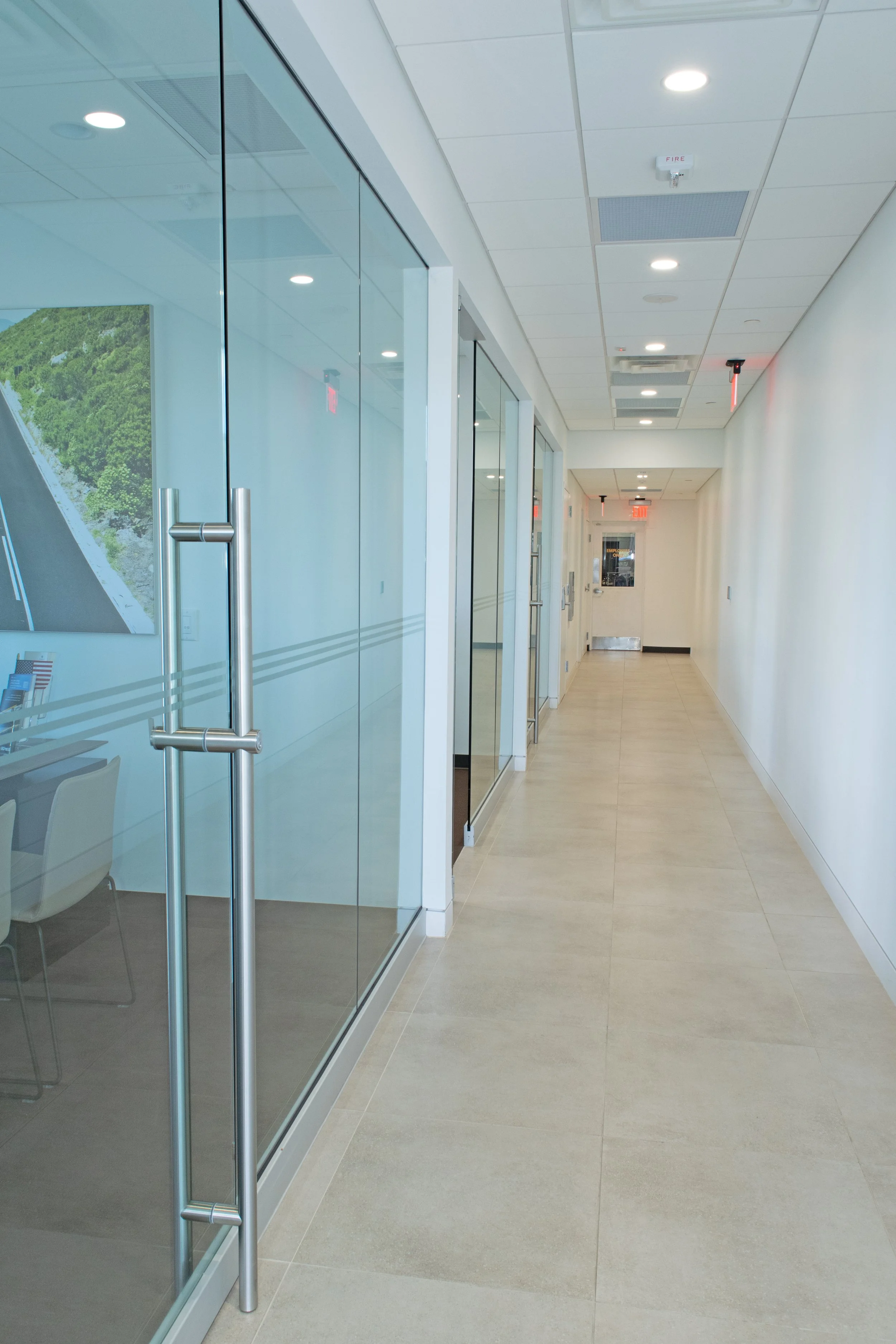 Modern office hallway with glass walls, metal handles, ceiling lights, and a fire alarm on the ceiling.