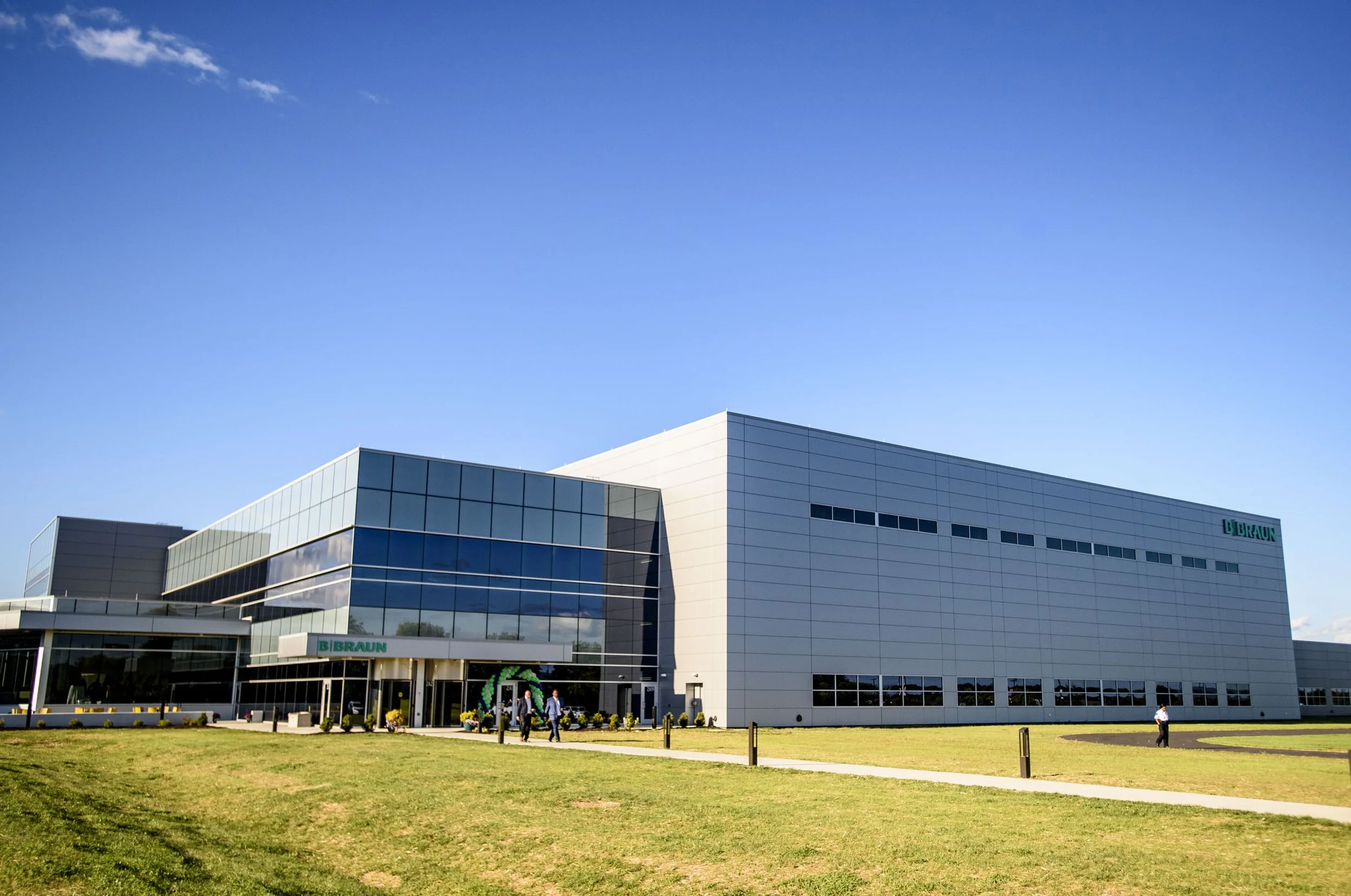 Modern office building with glass windows and white panels, labeled 'B. Braun,' with a green logo, under a blue sky with a few clouds. People are walking on a sidewalk in front of the building, and the area has a grassy lawn.