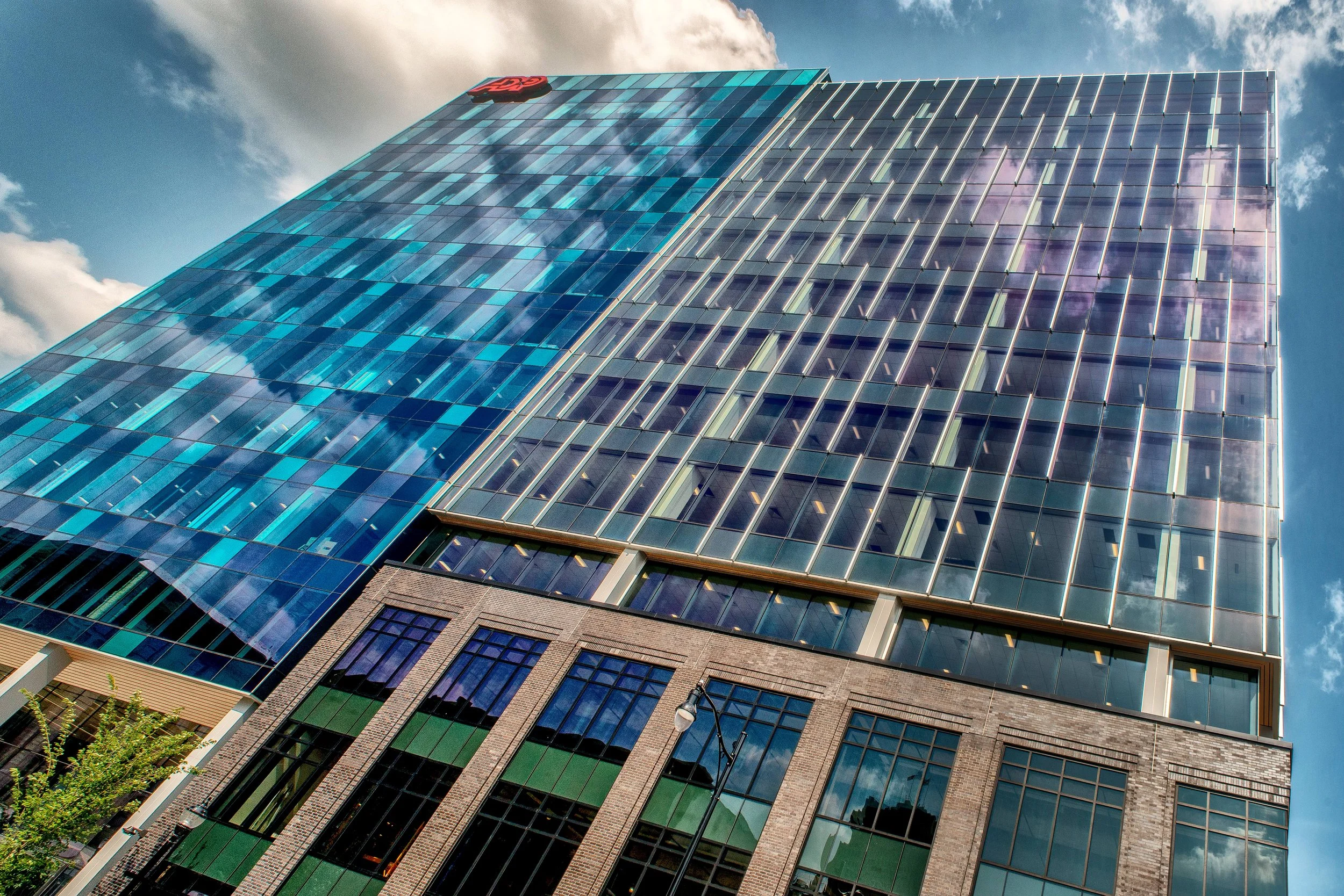 A tall modern glass skyscraper reflecting the sky and clouds, with a red logo at the top.
