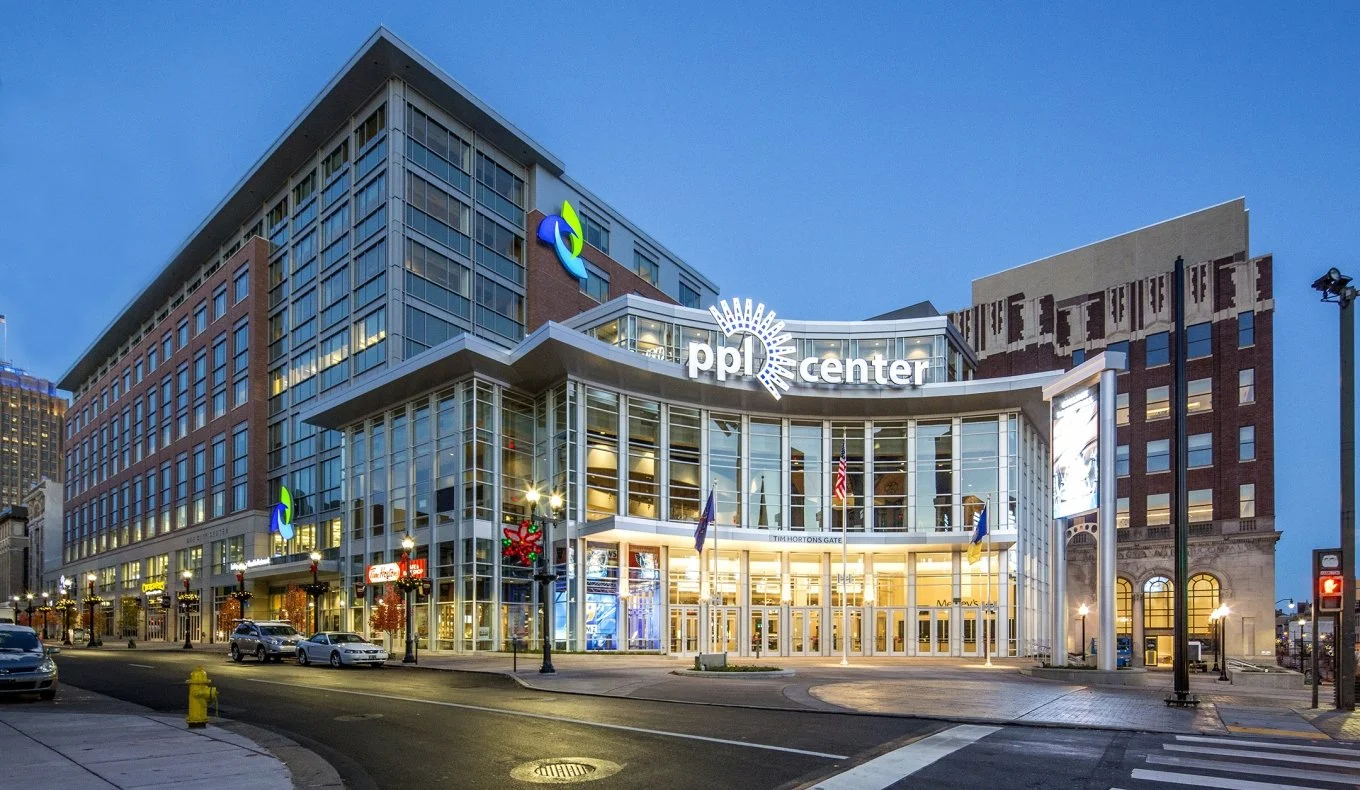 Exterior view of the PPL Center, a modern glass-fronted building with shopping and entertainment amenities, city street in the foreground, some cars, signage, and holiday decorations, during dusk.