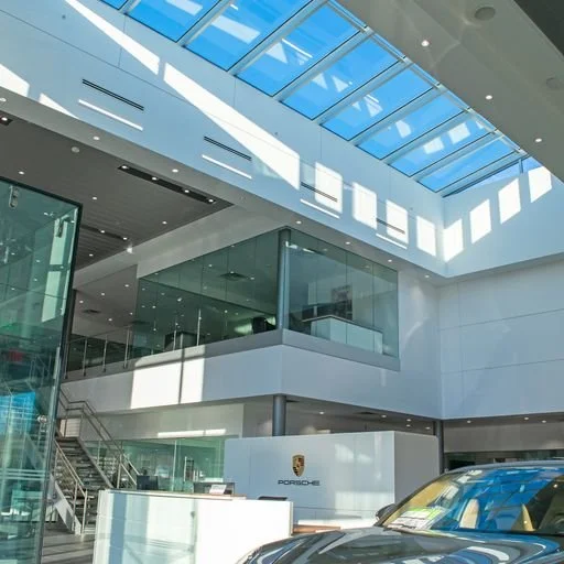Modern car showroom with a Porsche logo, glass walls, and a sleek yellow car visible at the front.