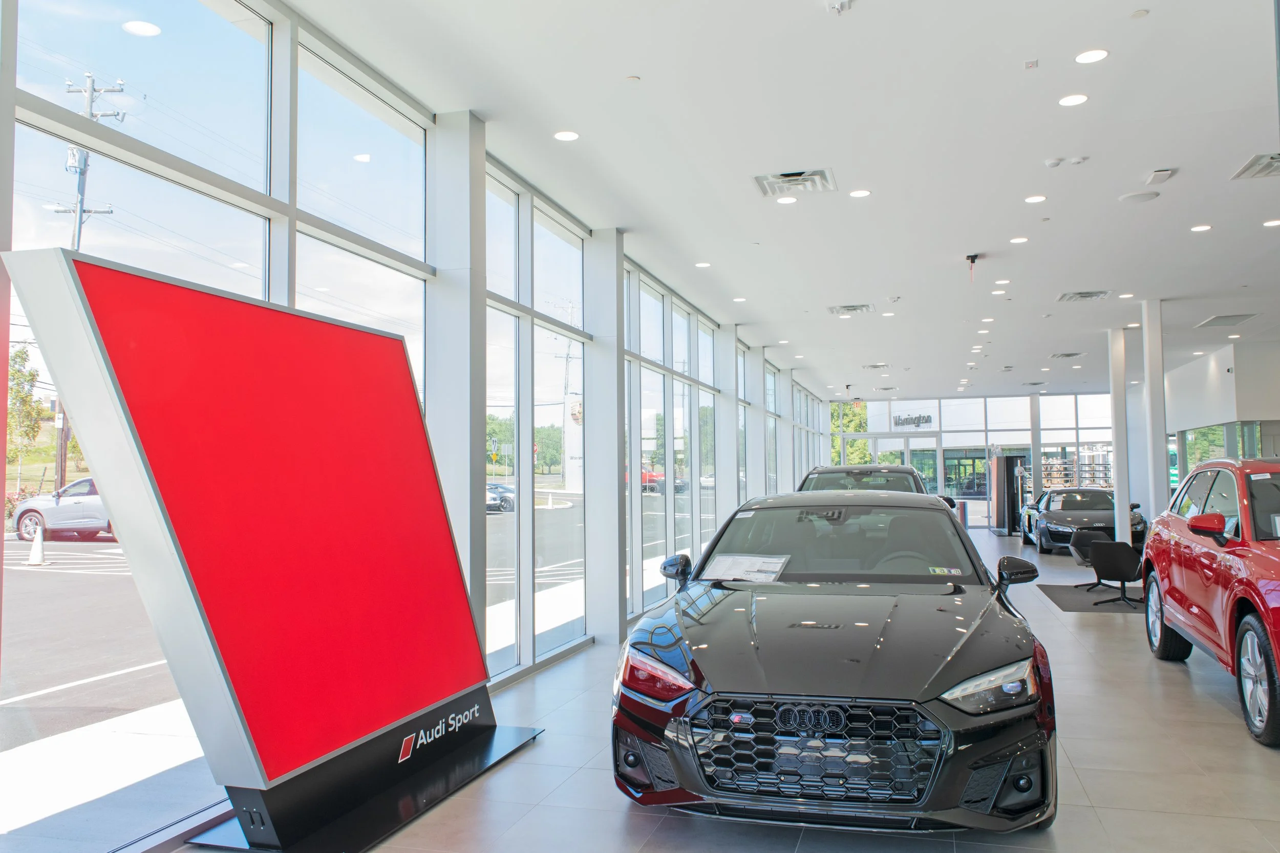Inside a Car dealership showing multiple new cars on display near large glass windows with a red promotional sign that says 'Audi Sport'.
