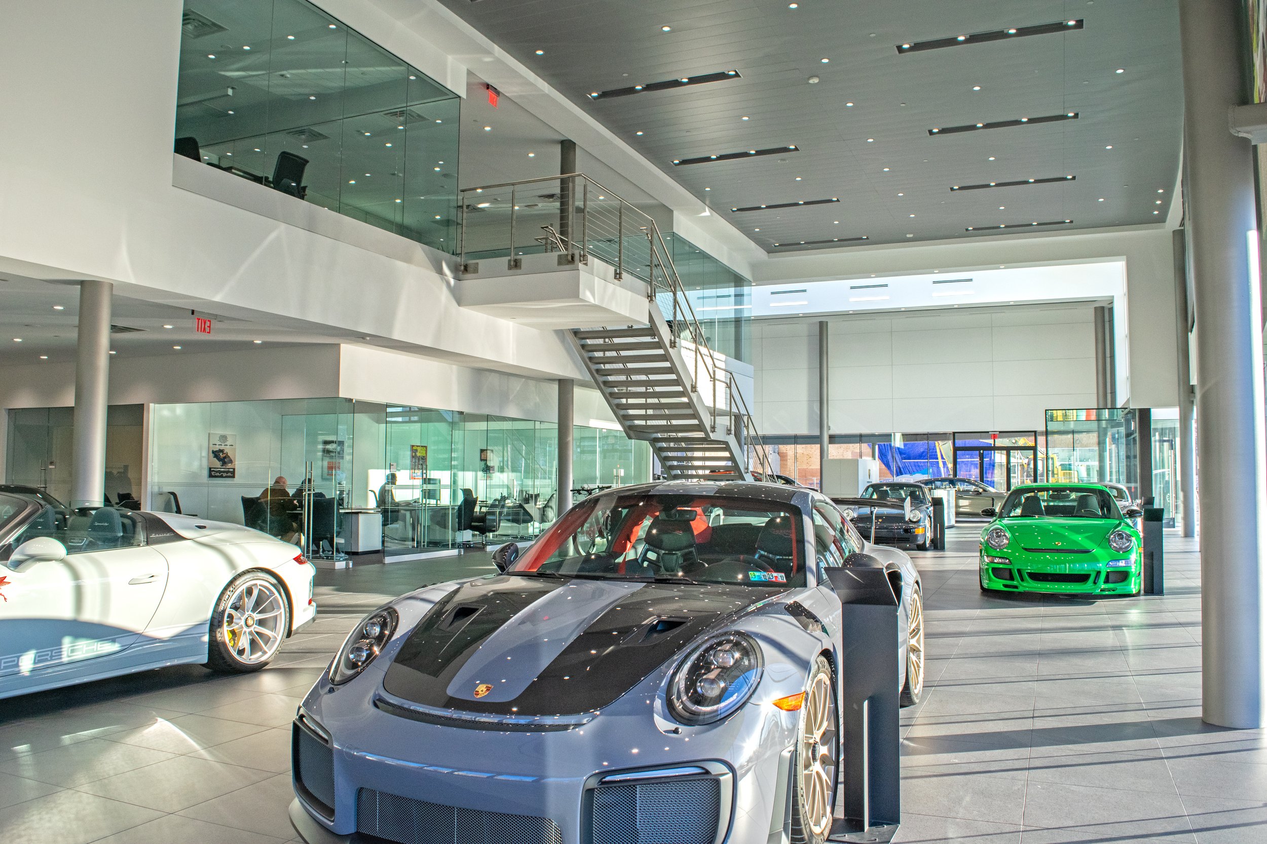 Showroom with luxury cars including Porsche and sports cars displayed indoors, with a modern, bright interior featuring glass walls, a staircase, and ceiling lights.
