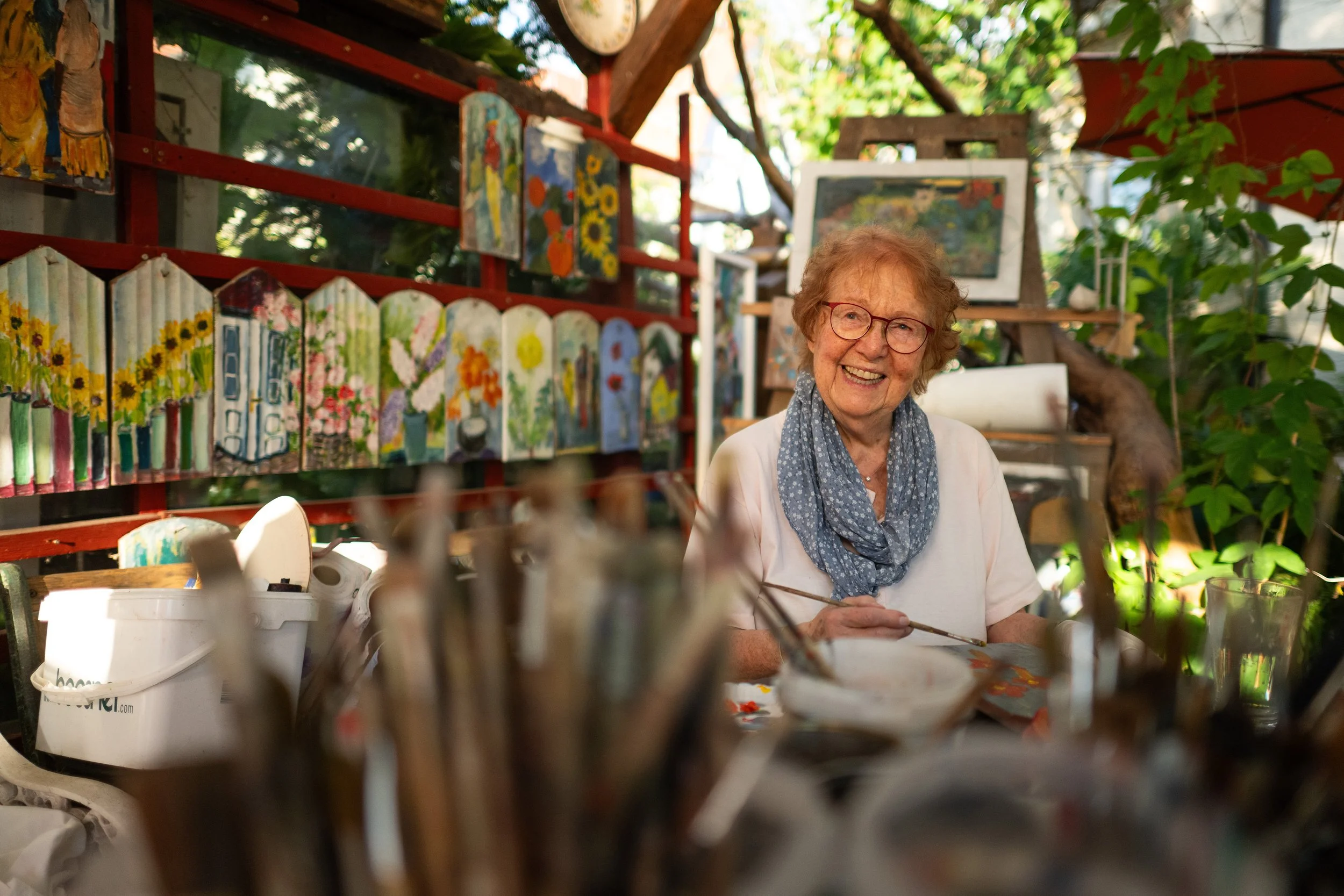 Eine ältere Frau mit roten Haaren, Brille und einem blauen Schal, die lächelnd in einer kunstvoll gestalteten Gartenlaube sitzt, umgeben von bunten Gemälden und Kunstwerken.
