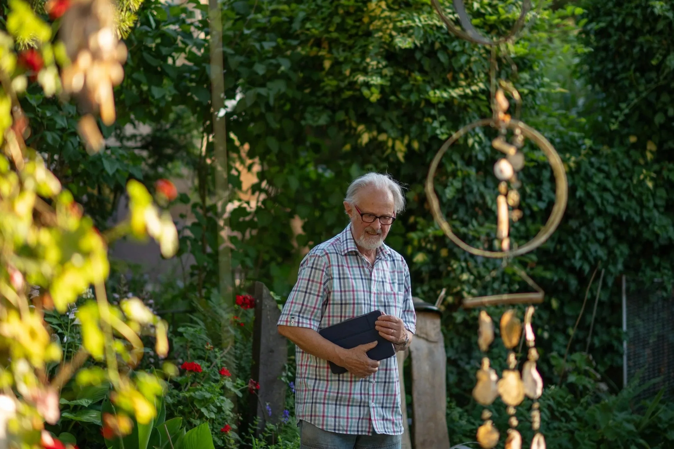 Ein älterer Mann mit Brille, grauem Haar und kariertem Hemd steht in einem Garten mit viel Grün und Blumen. Er hält ein schwarzes Tablet in den Händen und lächelt.