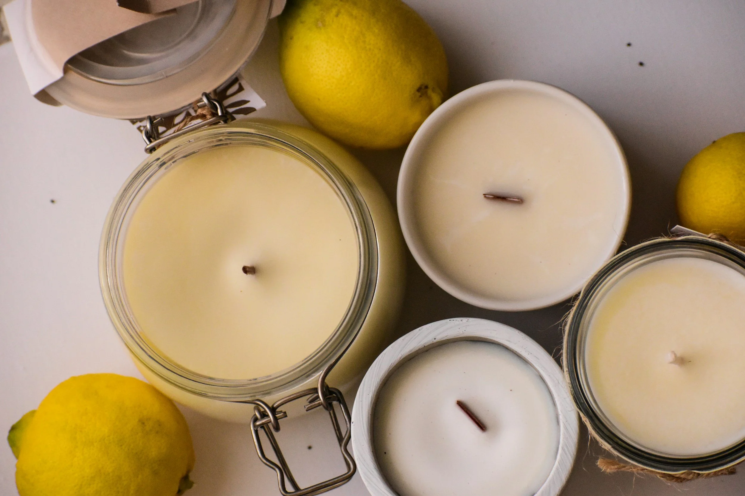 Candle jars with white wax and wicks, lemons, and a jar with a hinged lid on a white surface.