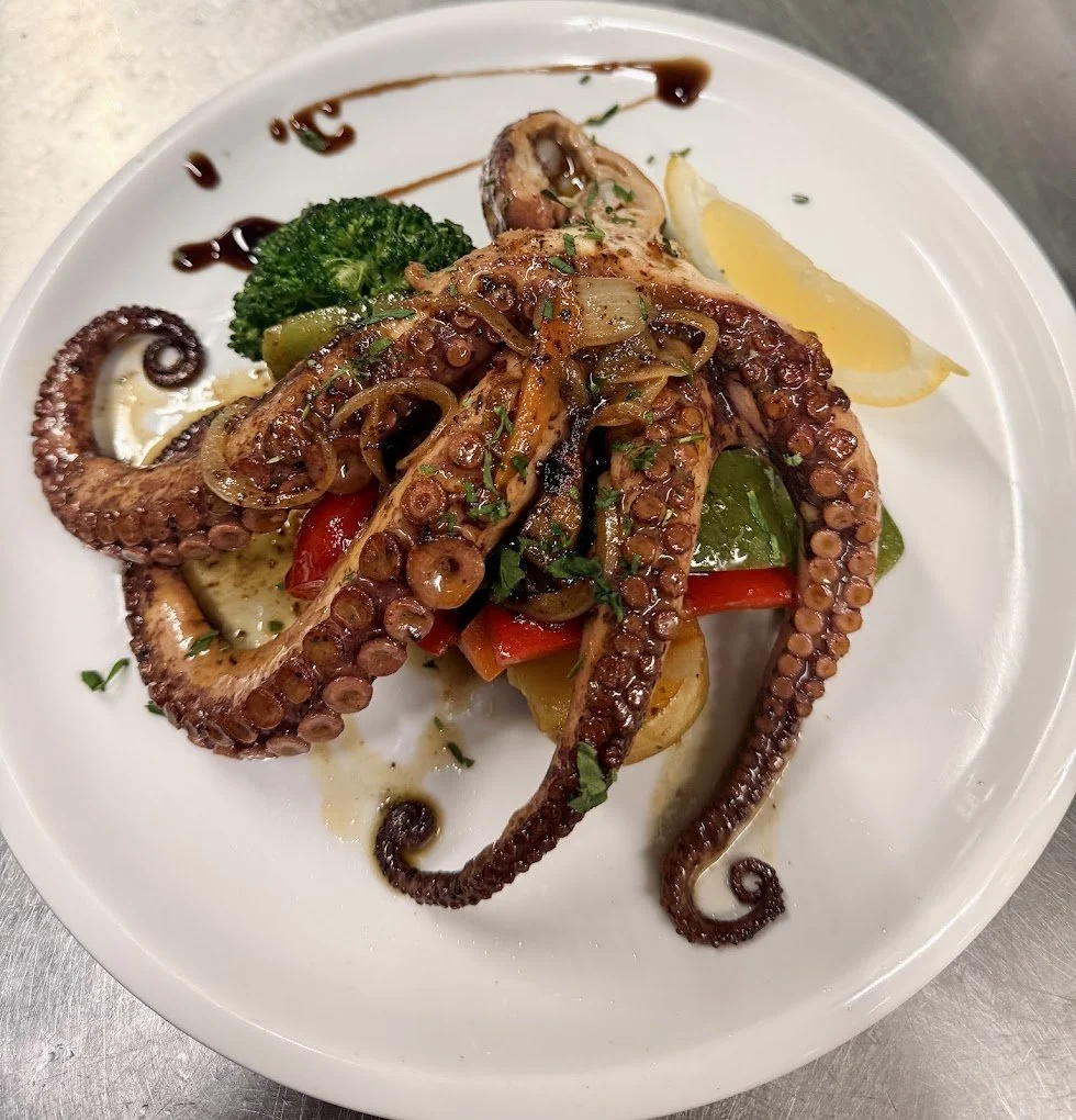 Plate of grilled octopus tentacles garnished with sautéed onions, herbs, vegetables, a lemon wedge, and broccoli.