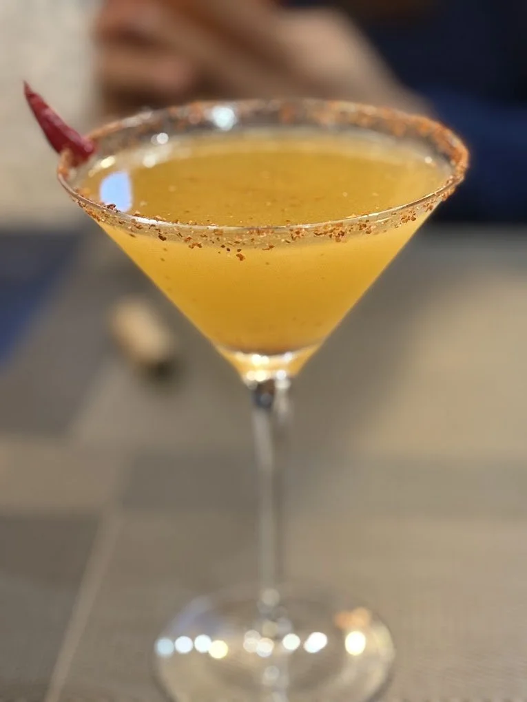 Yellow cocktail with a red chili garnish in a martini glass with a salted rim.