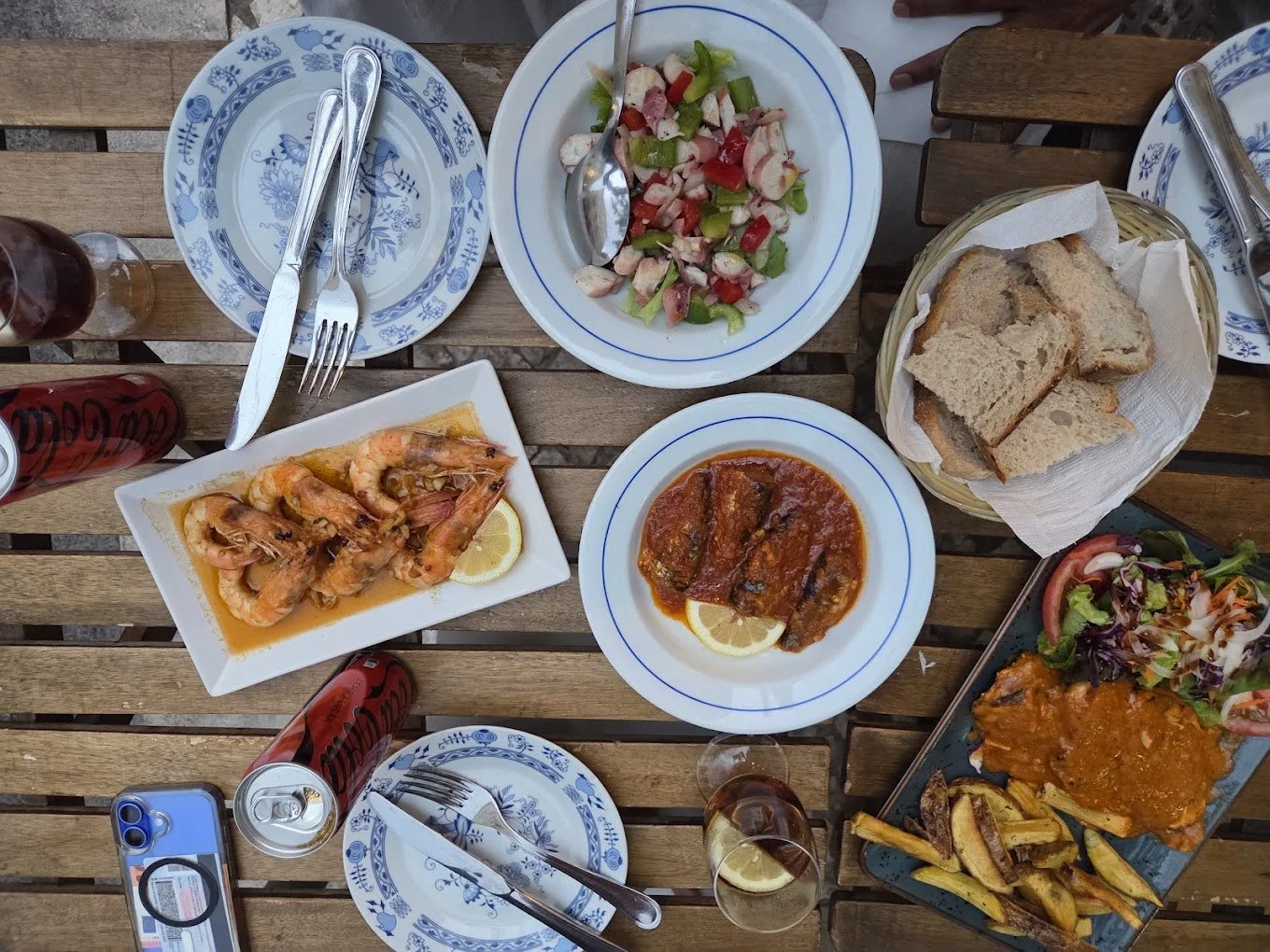 A wooden table set with plates of shrimp, salad, bread, stew, and fried plantains, with drinks and utensils.
