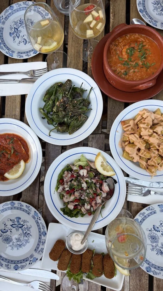 A table set with various dishes including a bowl of red soup, pasta salad, grilled green peppers, a vegetable salad with radishes, a plate of croquettes with dipping sauce, lemon slices, and glasses of white wine with fruit pieces, all on a rustic wo