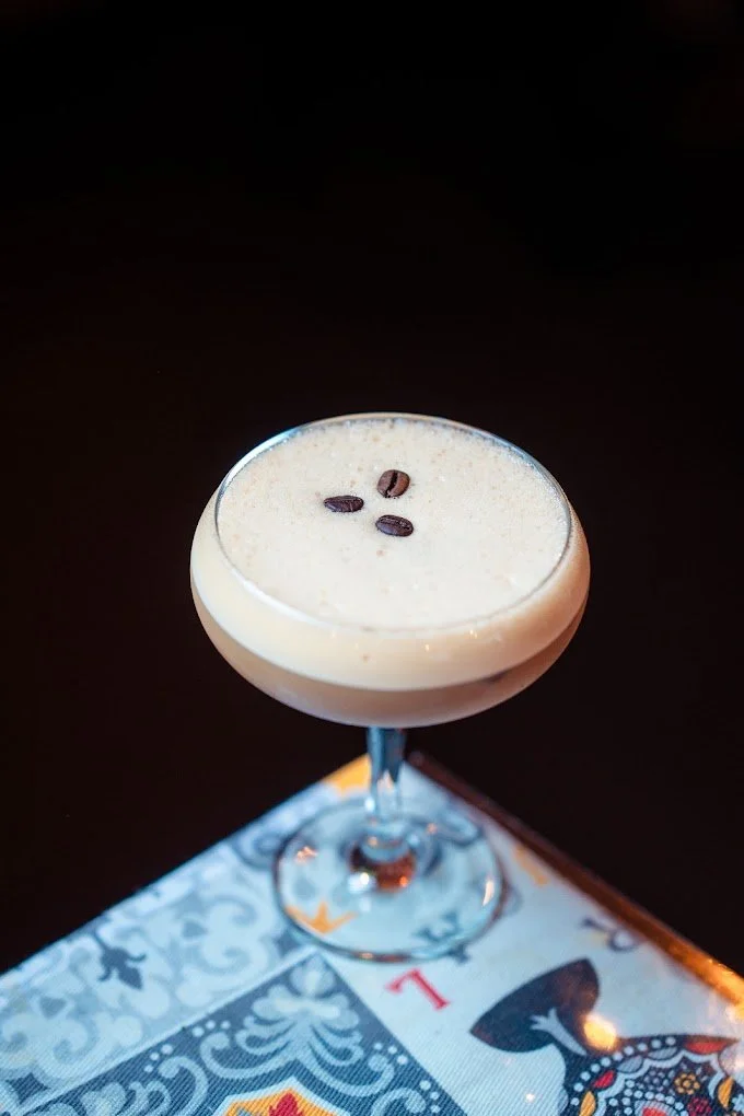 A creamy coffee drink garnished with coffee beans in a coupe glass on a decorative tablecloth.