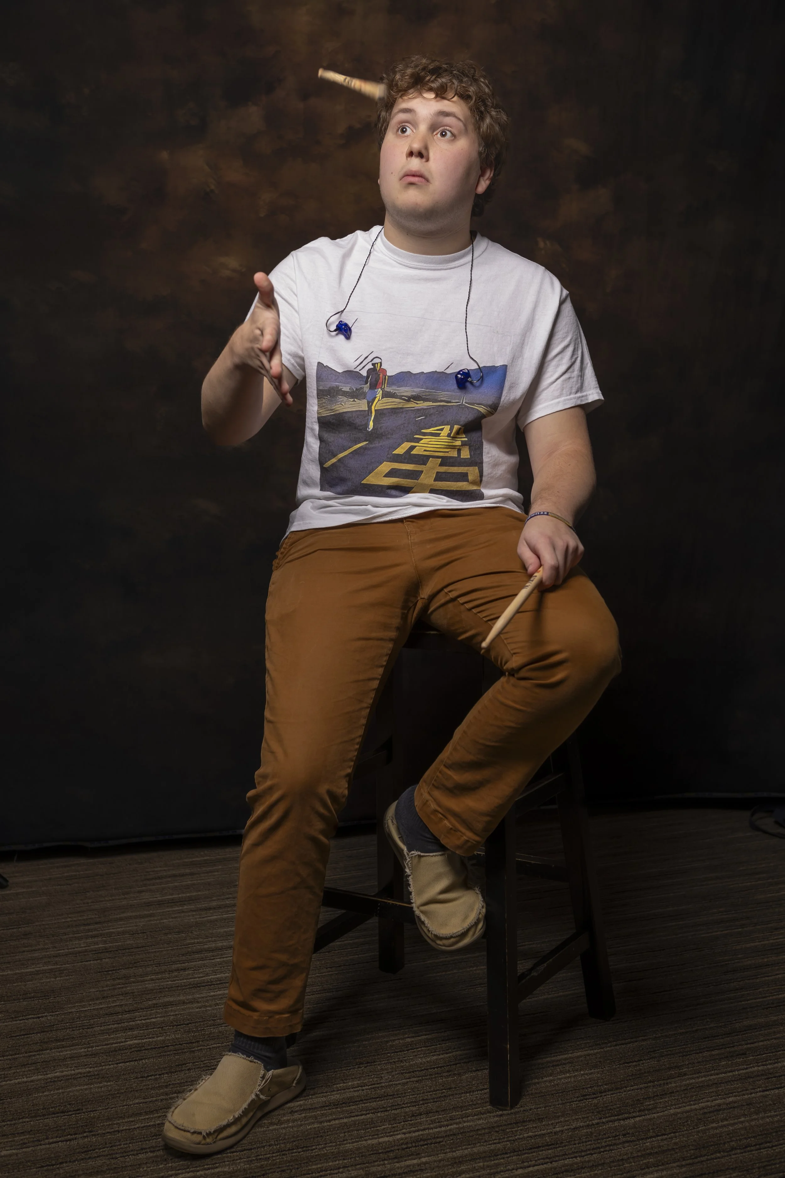 Young man with curly hair sitting on a black stool, wearing a graphic t-shirt, brown pants, and beige slip-on shoes, with a dark brown textured background.
