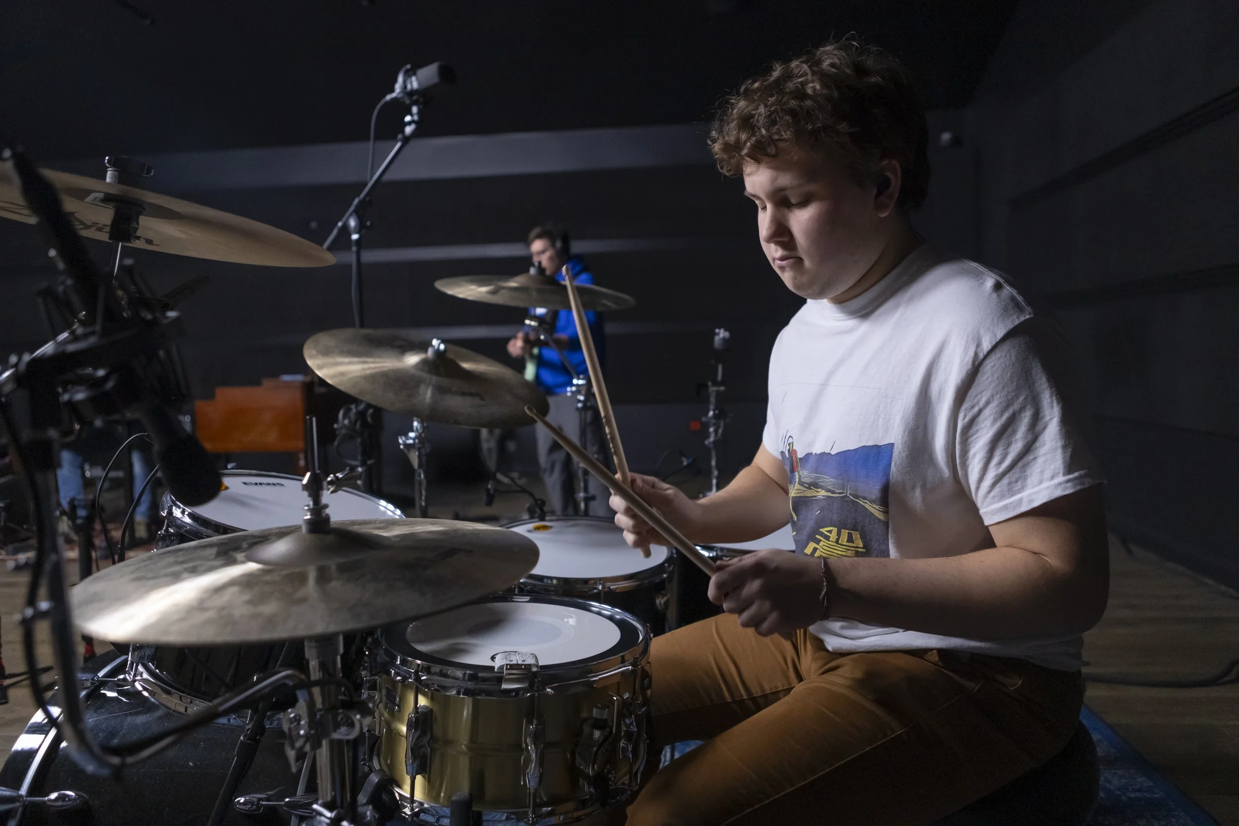 Young man playing drums in a dimly lit music studio, with another musician in the background.