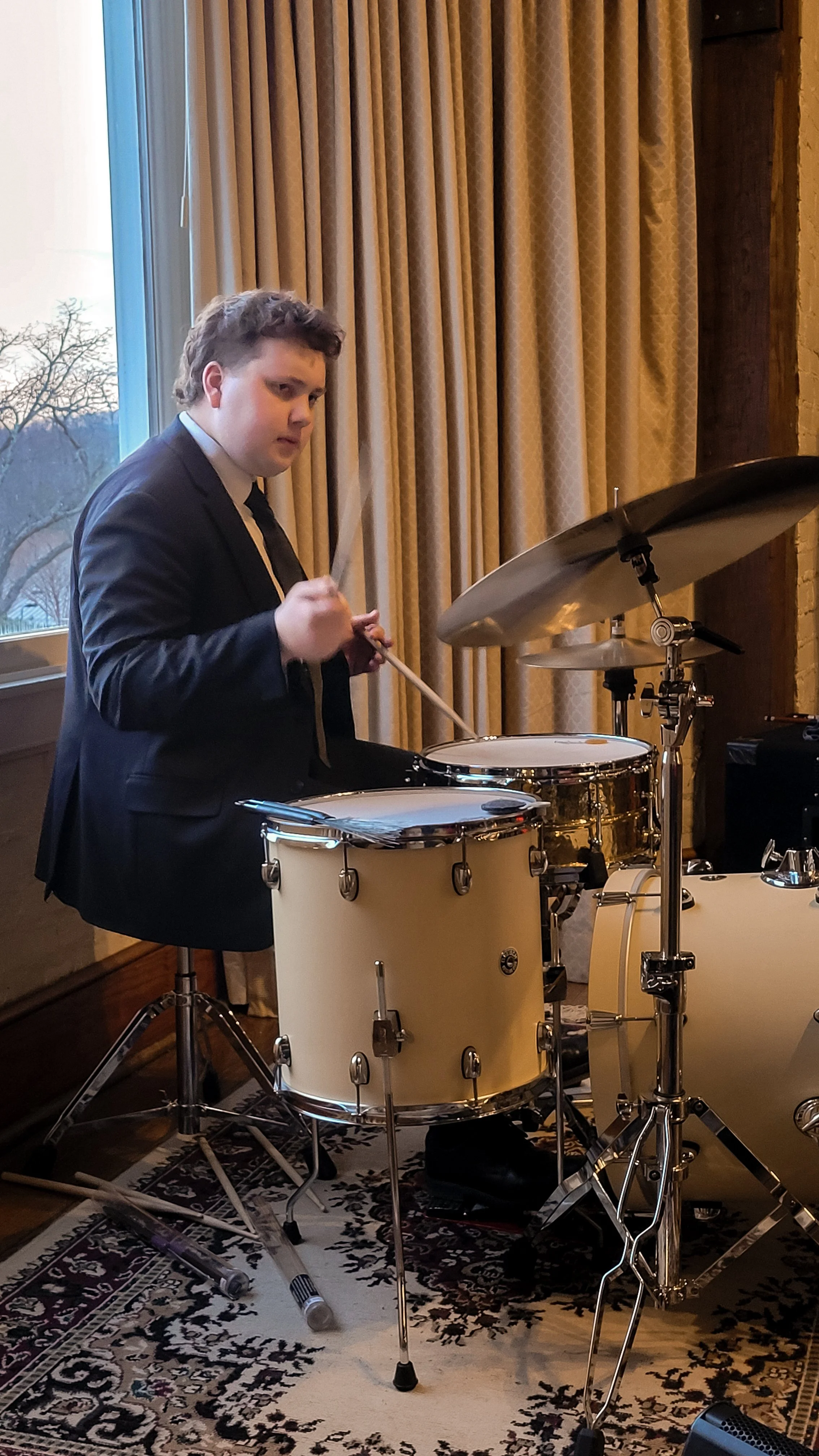 Young man in a suit playing a drum set indoors next to a window with beige curtains, with a patterned rug underneath.