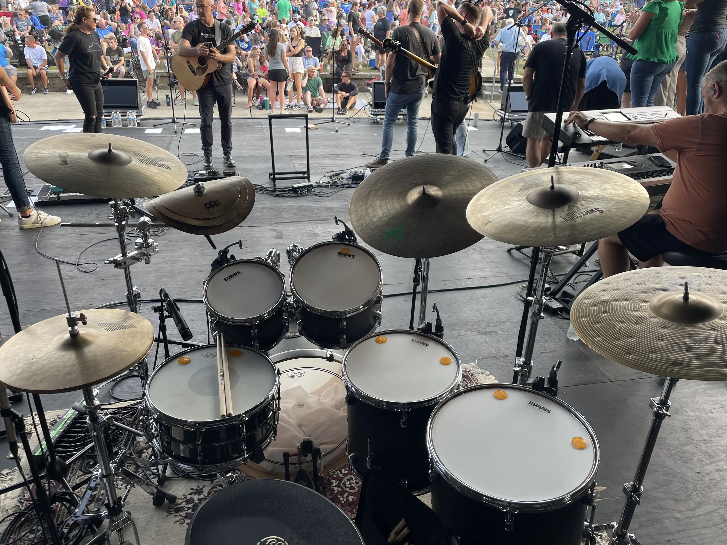 Stage setup with drums, keyboards, and microphones before a live outdoor concert. Audience seated in the background, some standing and socializing.