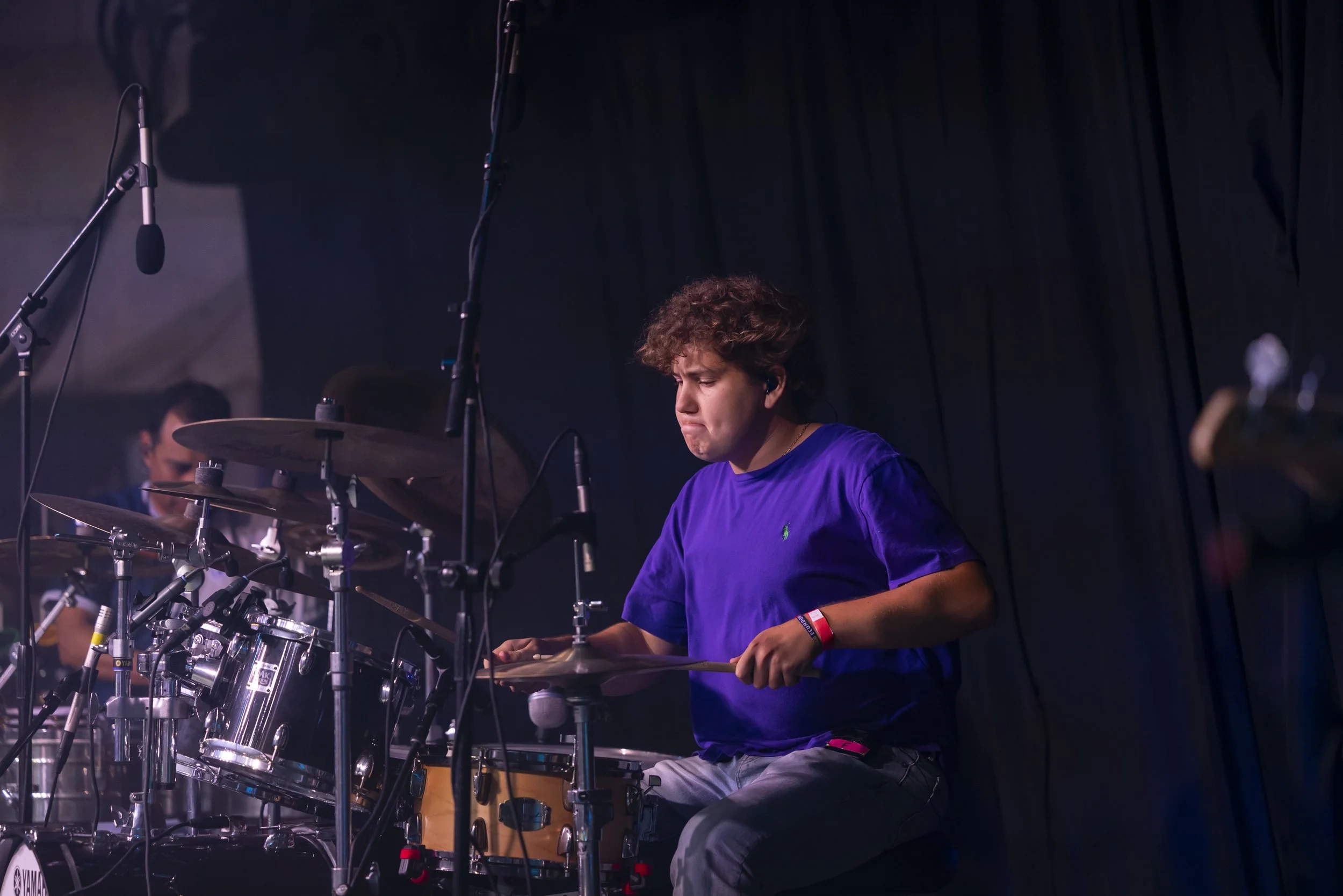A young person with curly hair playing drums on stage during a performance.