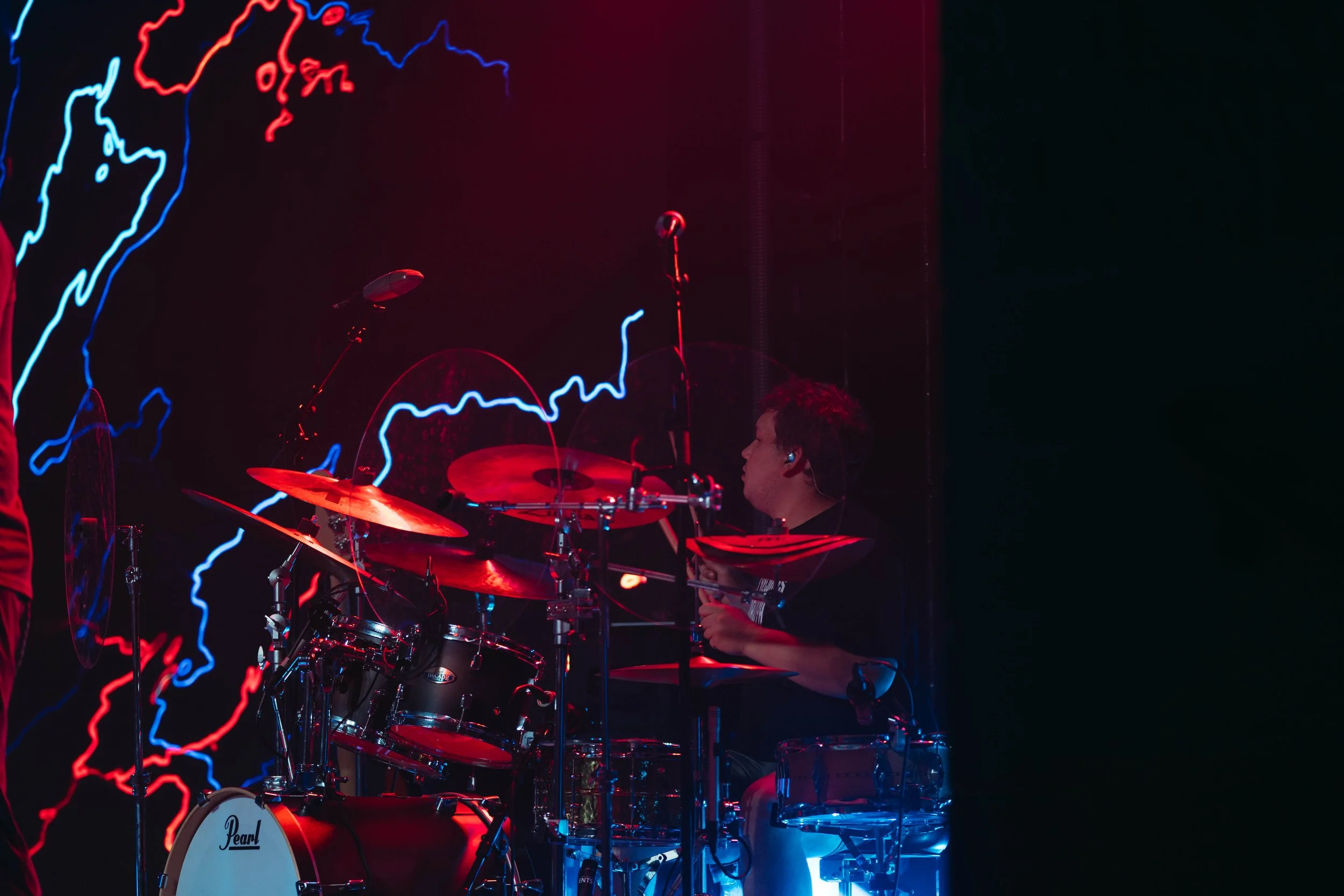 A young drummer playing a drum set on stage with neon light art of a map behind him.