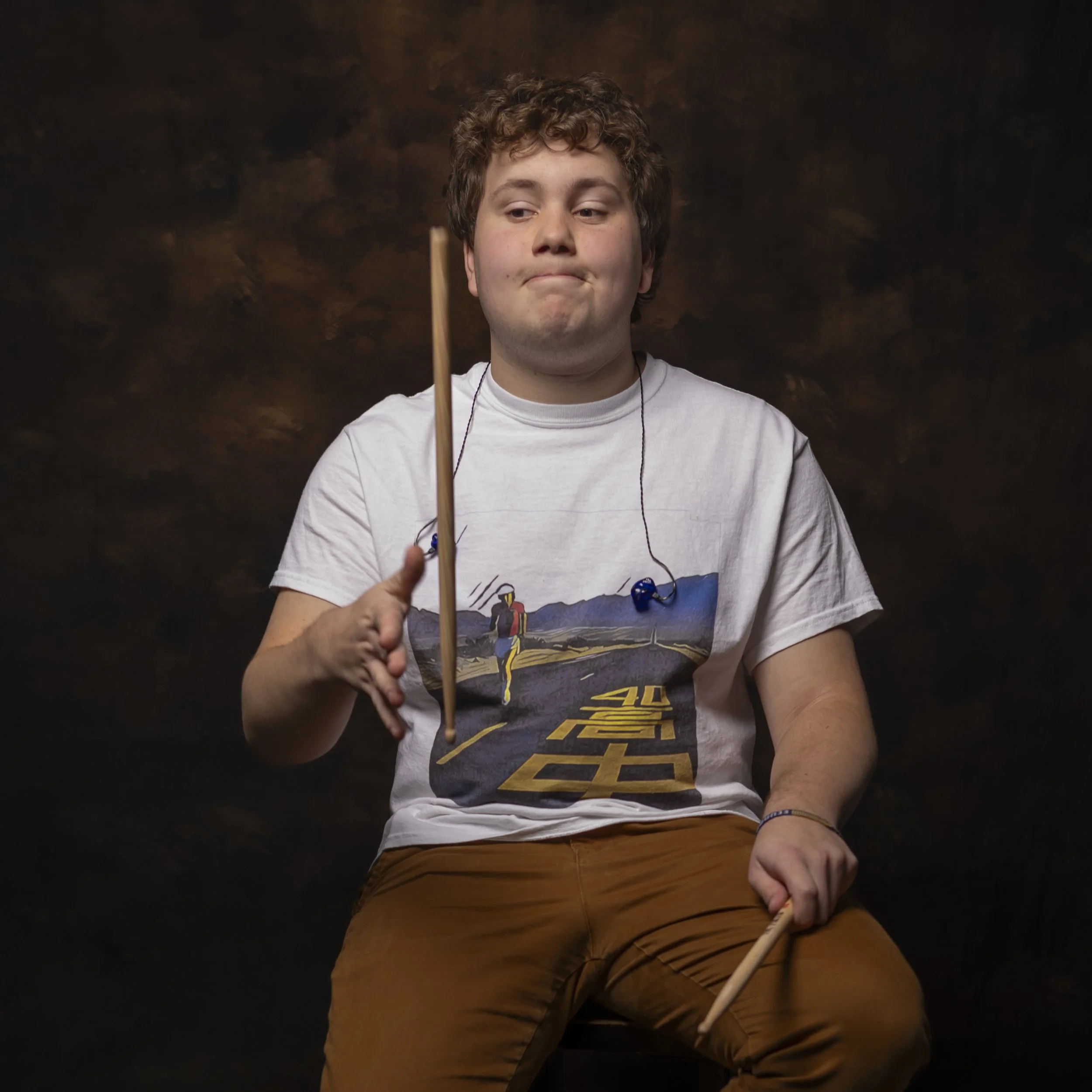 A young man with curly hair wearing a white T-shirt and brown pants sitting against a dark brown textured background, holding a drumstick in his right hand.
