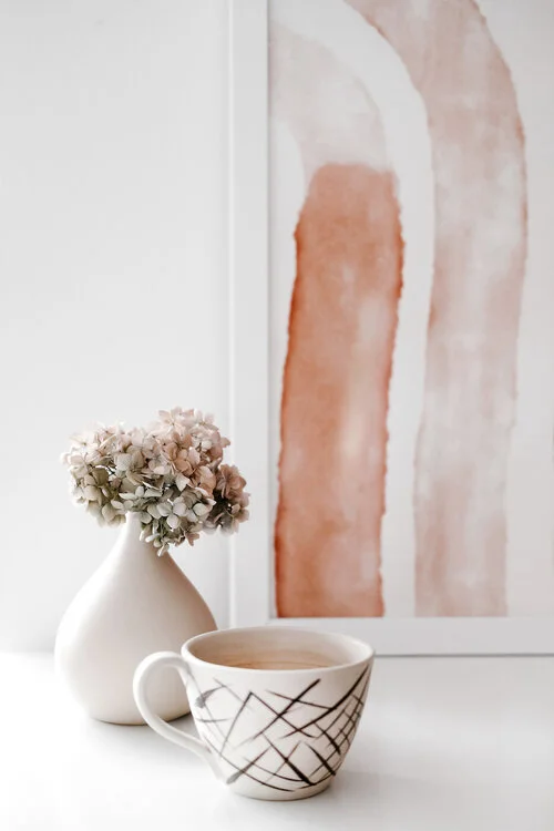 A white vase with dried hydrangea flowers, a ceramic mug with black lines, and a framed abstract painting with earth tones on a white surface.