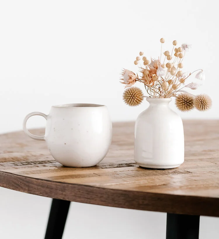 A white ceramic mug and a small white vase with dried flowers on a wooden table.
