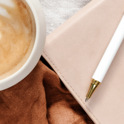 Close-up of a beige notebook, white pen with gold accents, and a cup of coffee on a brown and beige cloth surface.
